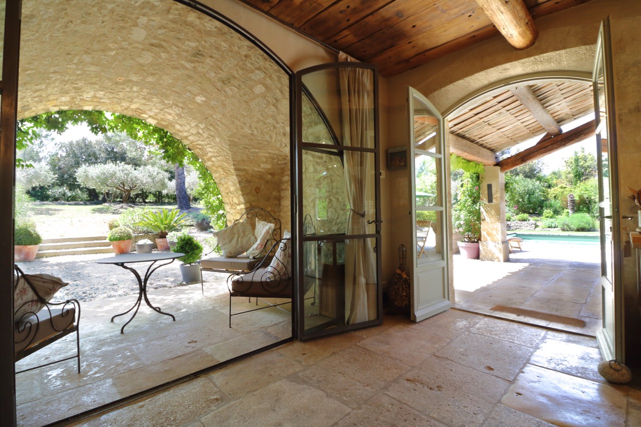 Patio doors open to a tranquil garden from an arched, stone-floored veranda with wooden beams, featuring a comfortable seating area with armchairs and a chaise lounge, inviting a relaxing outdoor experience.